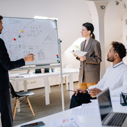 Responsable de equipo explicando objetivos en pizarra a mandos intermedios en sesión de liderazgo.